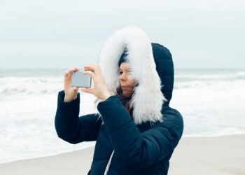 Young woman in a winter jacket with a hood using a mobile phone - Meteo Italia: fine del maltempo grazie all’alta pressione