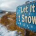 A rustic wooden sign with the words “Let It Snow” painted in white against a blue background with snowflakes, set against a backdrop of a snowy road and a snowy landscape. - Meteo: nuovo ciclone colpirà duramente queste regioni