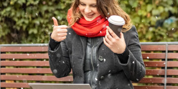 Young woman working on laptop sitting on a park bench with coffee. Computer and online shopping use. Distance learning, online education. - DIRETTA METEO, News Meteo, Previsioni Meteo, Viaggi