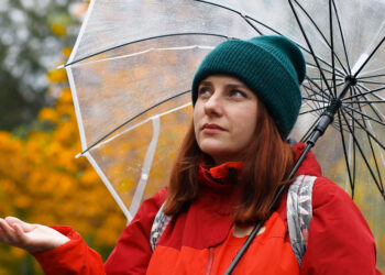 Young girl in warm clothes with a transparent umbrella and a backpack on her back outside in cold weather after rain. Woman looks at the camera. - Meteo: forte ondata artica in arrivo in Italia, ecco dove nevicherà