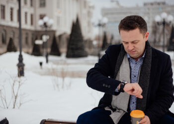 A young man in a coat and suit sits on a bench in a city park looking at his watch with a cup of coffee - Forse nevicherà per l’Immacolata?