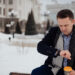 A young man in a coat and suit sits on a bench in a city park looking at his watch with a cup of coffee - Meteo, arriva un nucleo gelido: ecco cosa aspetta l’Italia