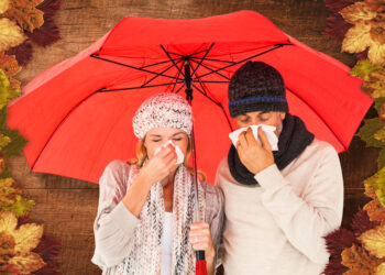 Ill couple sneezing in tissue while standing under umbrella against overhead of wooden planks - Meteo: temperature in crescita in tutto il Paese, ma non dureranno