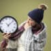 scared young ill girl wearing white robe and winter hat with scarf holding and looking at wall clock wrapped in plaid isolated on olive green background - Dicembre, intensa ondata artica ma anche rischio anticiclone meteo